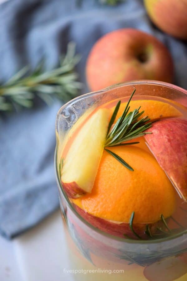 A glass pitcher filled with autumn harvest punch drink, featuring apple slices, orange, and rosemary.